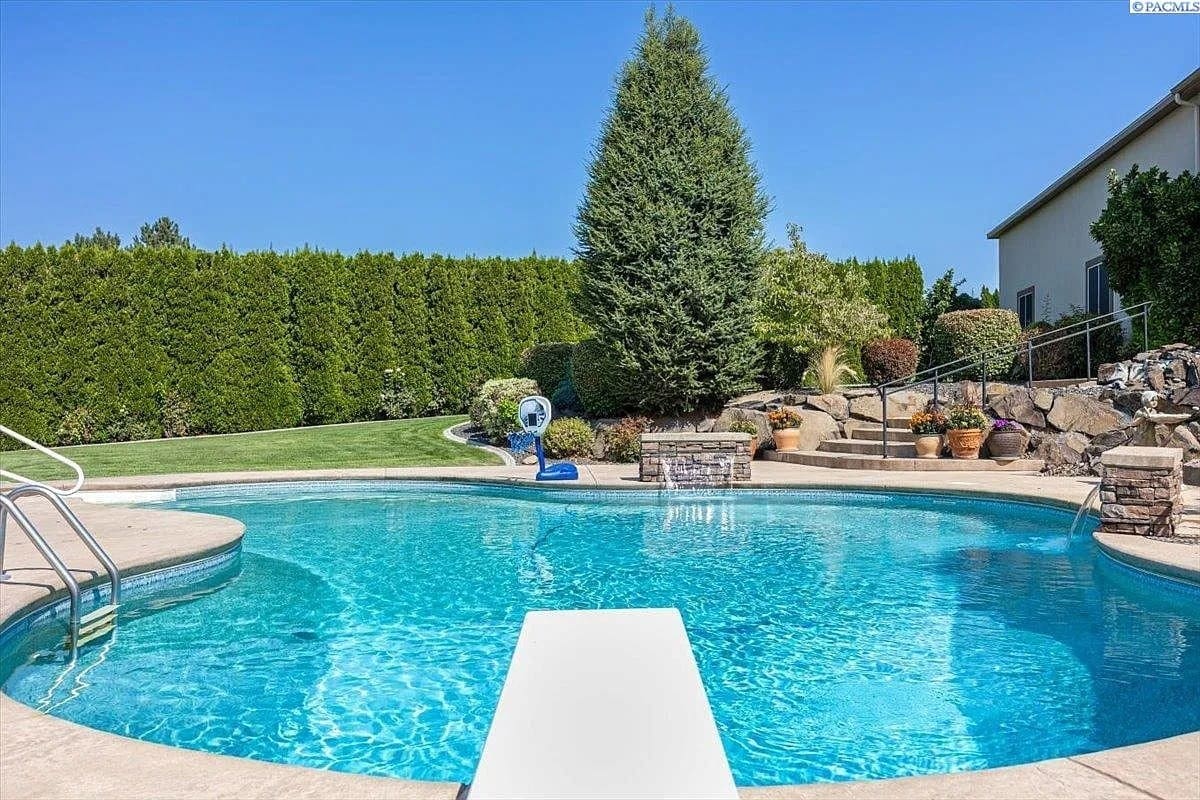 Backyard with a clear swimming pool, diving board, and surrounding greenery under a blue sky.