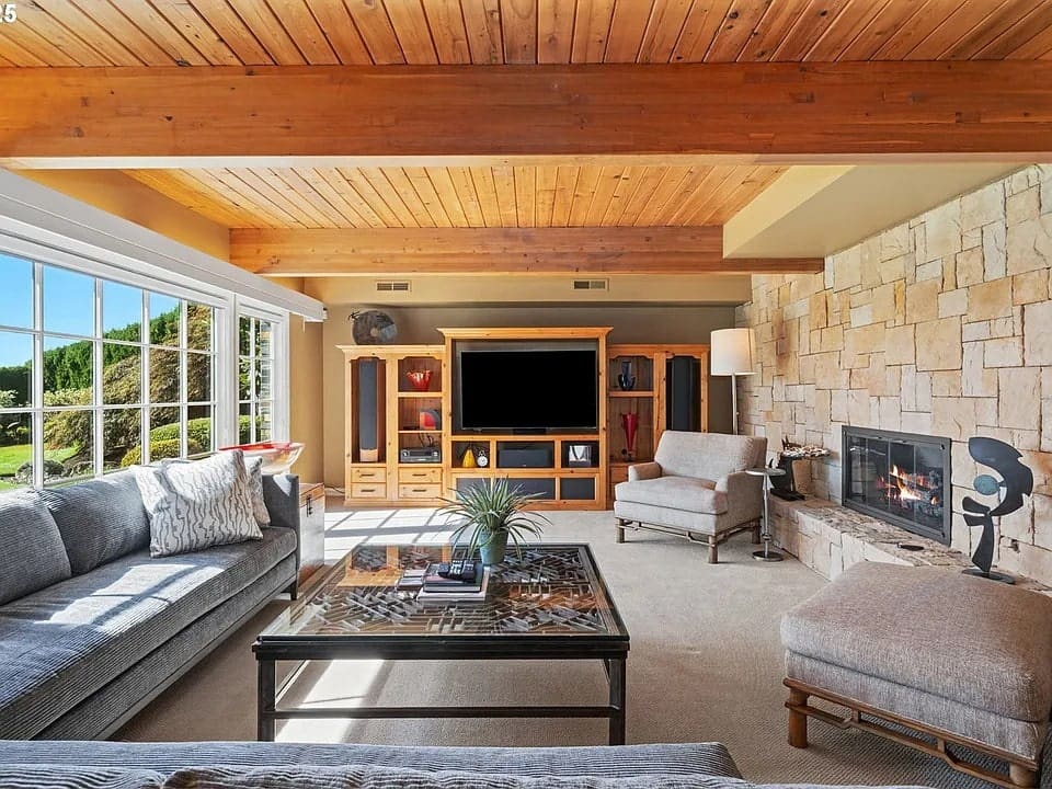 Modern living room with wooden ceiling, stone fireplace, large TV, and glass coffee table.