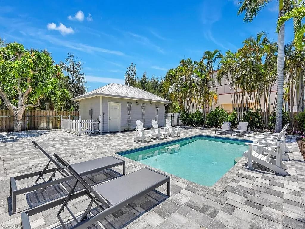 Outdoor pool area with lounge chairs, a small building, and surrounding palm trees under a clear sky.