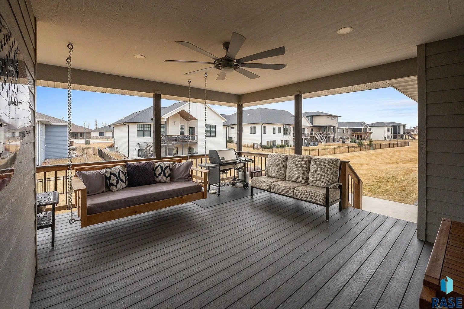 Covered patio with a ceiling fan, swing, sofa, and grill, overlooking a suburban backyard.