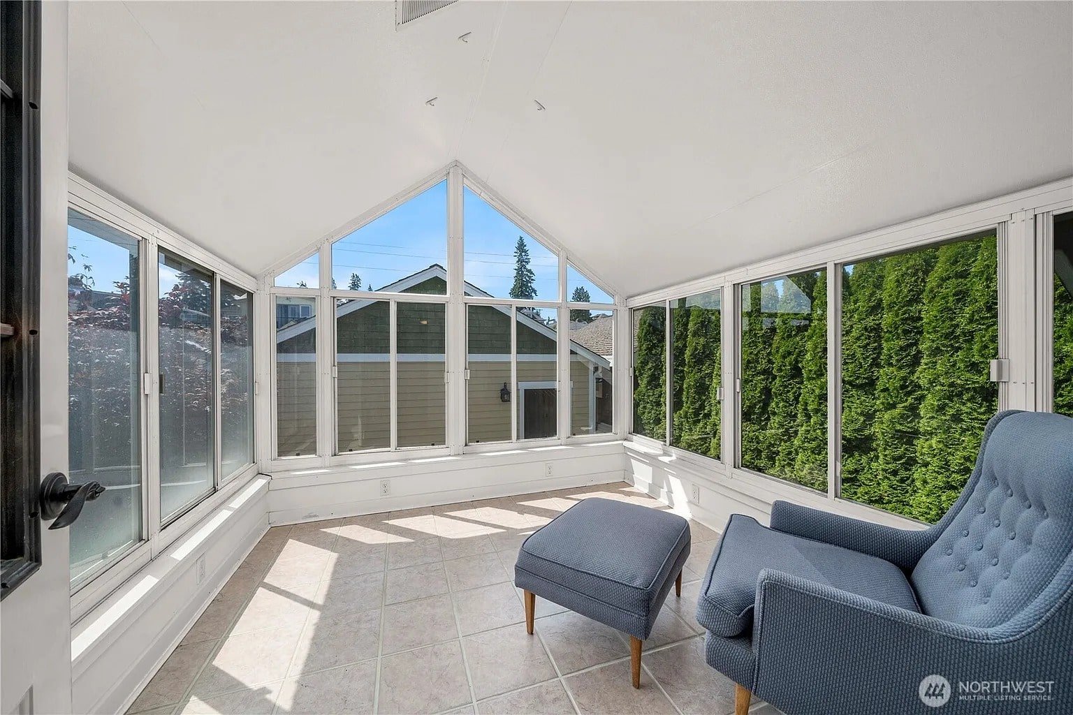 Sunlit room with large windows, blue armchair, ottoman, and tiled floor, overlooking greenery.