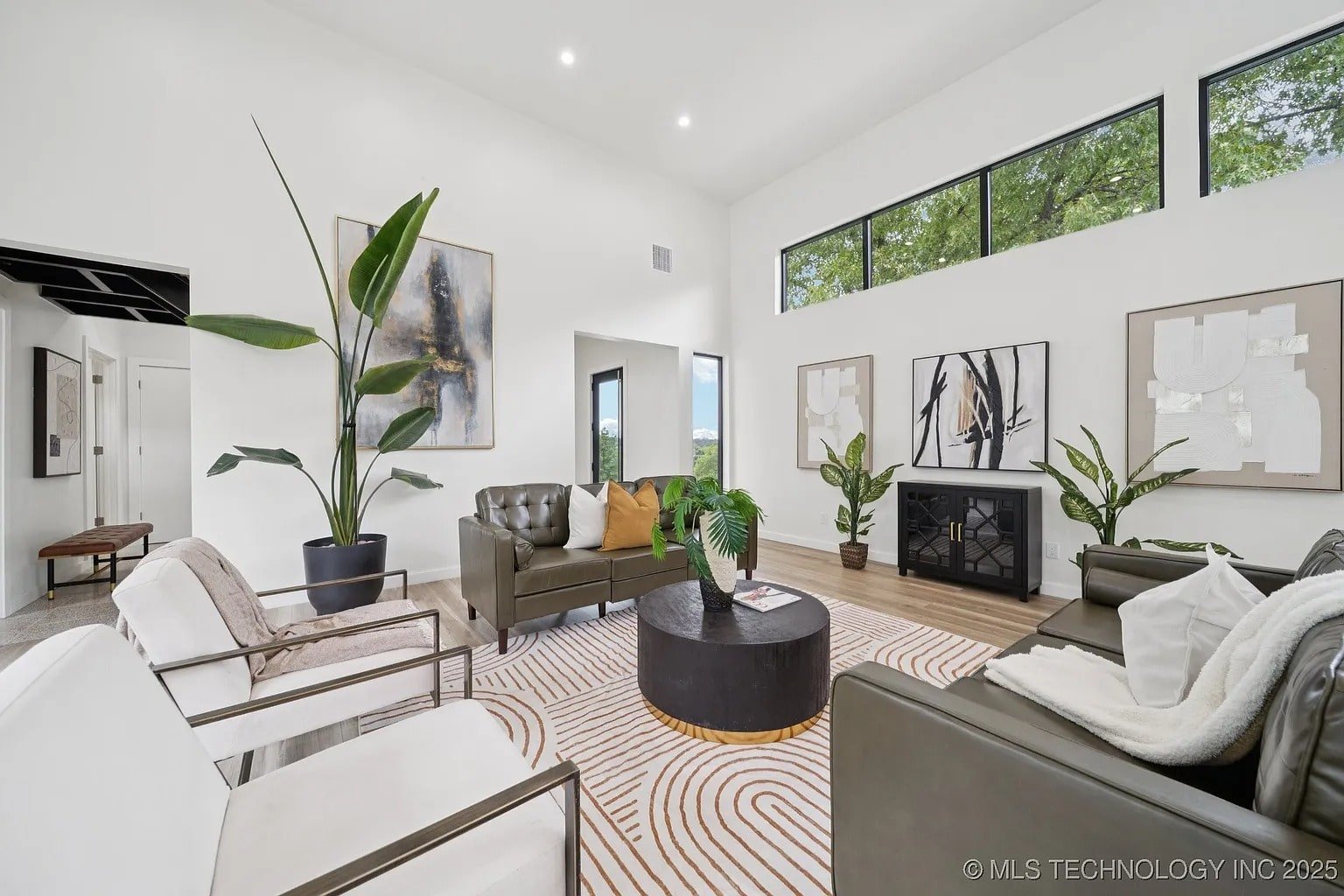 Modern living room with white walls, green sofas, abstract art, and large plants. Bright natural light.