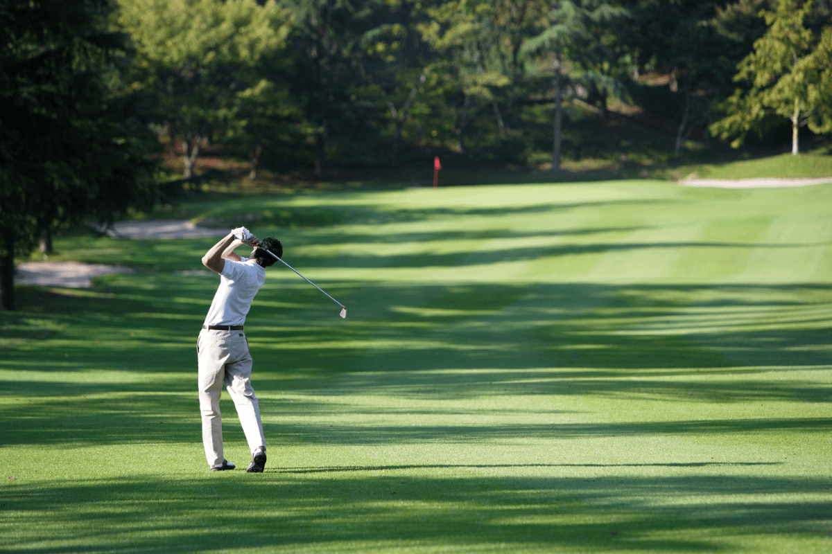 A golfer in a white shirt and beige pants swings a club on a lush, sunlit golf course with a red flag in the distance.