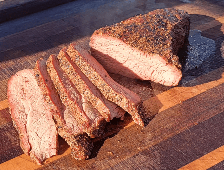 Sliced, seasoned smoked meat on a wooden cutting board, showing a pink interior.