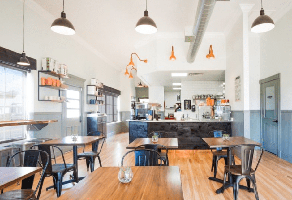 Modern cafe interior with wooden tables, metal chairs, and a counter with hanging orange lights.