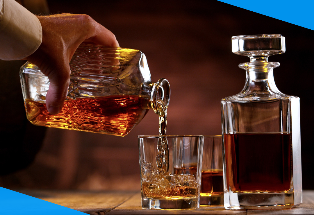A hand pours whiskey from a decanter into a glass with ice, next to another full decanter.