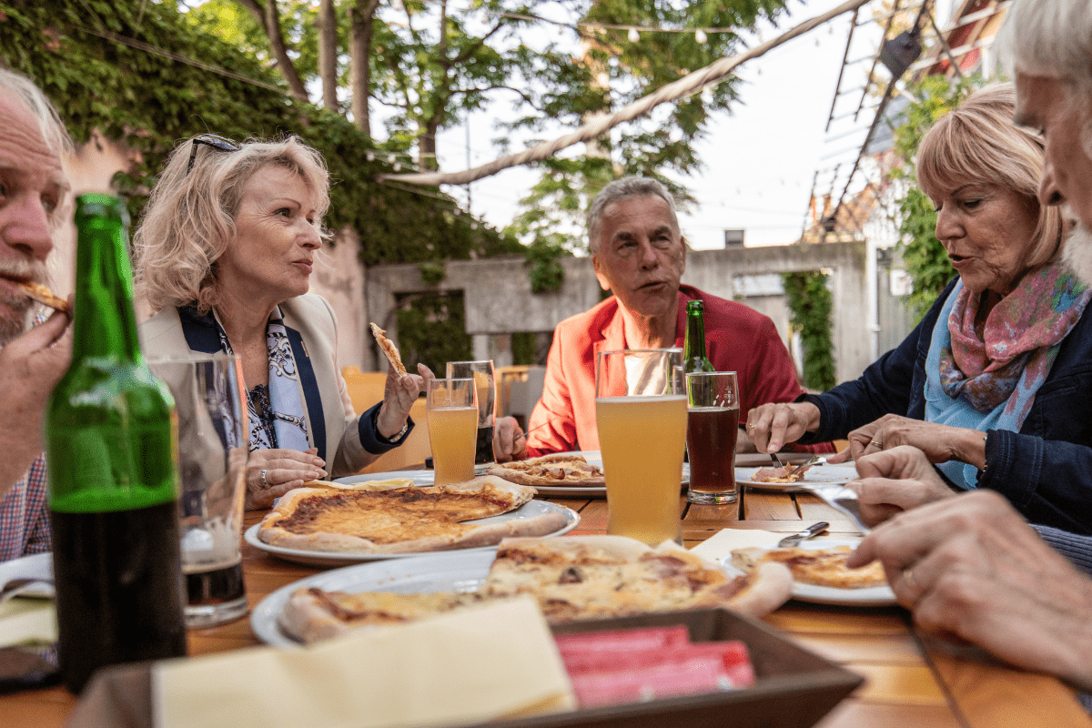 A group of older adults enjoying pizza and drinks at an outdoor table surrounded by greenery, with a variety of beverages and pizzas in front of them.
