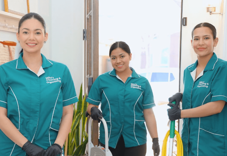 Three smiling cleaning staff in teal uniforms, holding cleaning equipment, standing indoors.