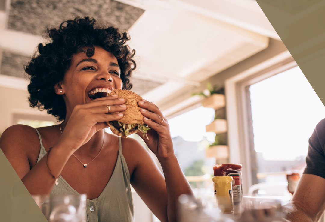 Woman joyfully eating a sandwich at a sunlit dining table.
