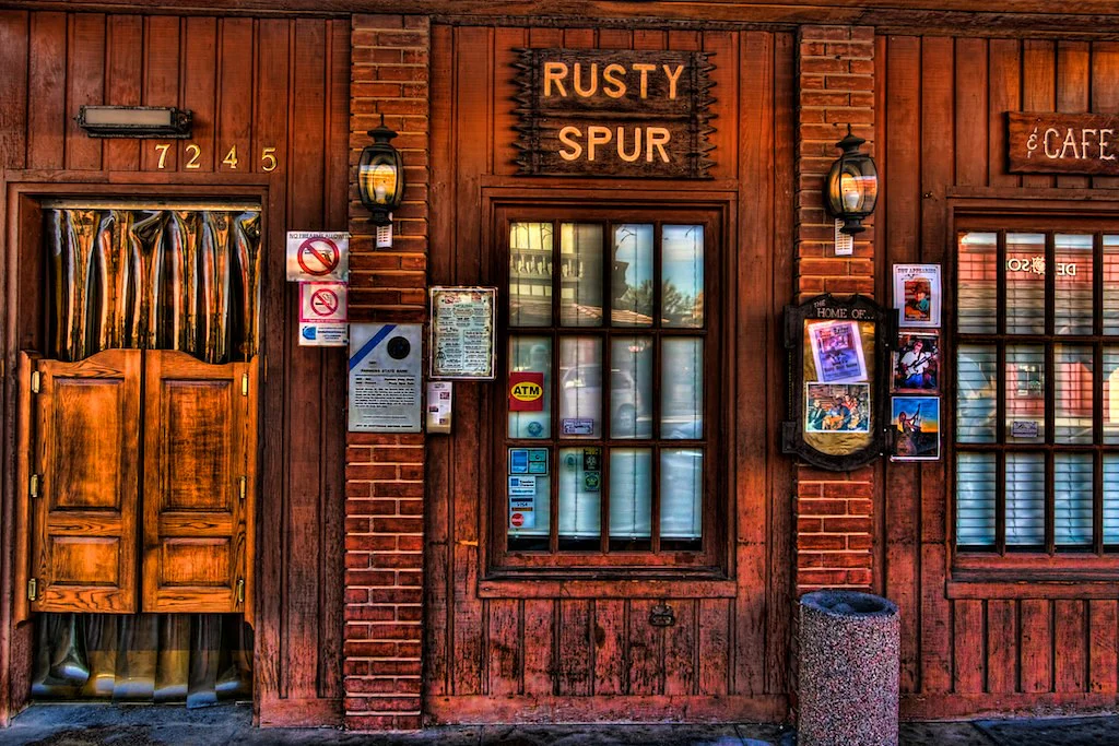 Wooden storefront with signs reading 'Rusty Spur' and 'Cafe'. Features a door, windows, and posters.
