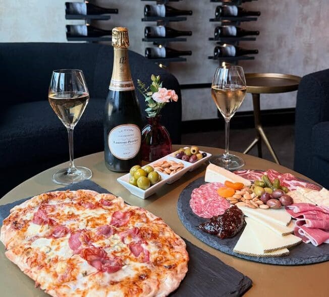 Pizza, charcuterie board, olives, and champagne on a table with wine glasses and wall-mounted bottles.