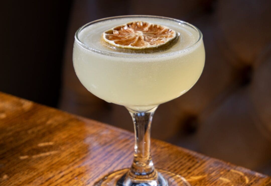 A cocktail in a coupe glass garnished with a dried citrus slice on a wooden table.