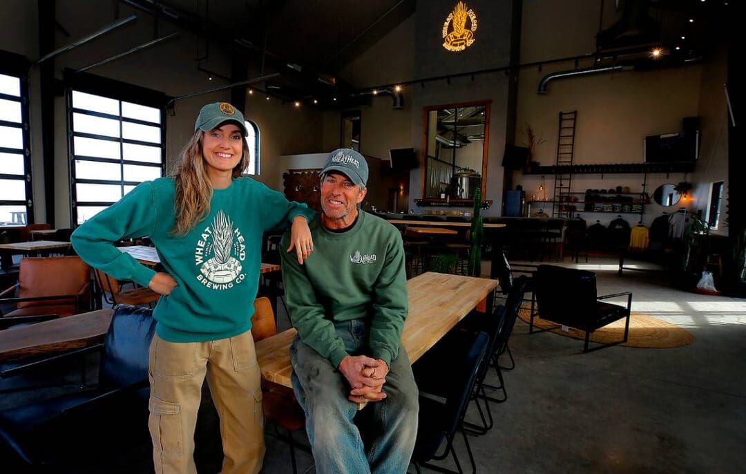 Two people smiling inside a brewery, wearing branded sweatshirts and hats.
