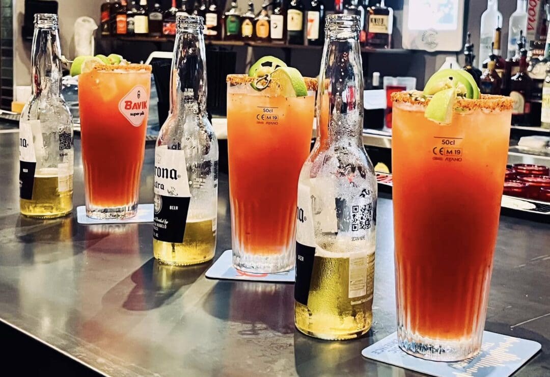 Five micheladas with lime and beer bottles on a bar counter.