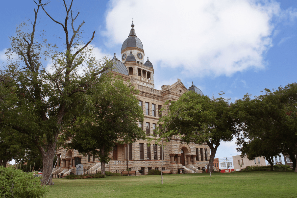 Historic stone courthouse with a central clock tower and ornate architectural details, surrounded by lush green trees and a clear blue sky.
