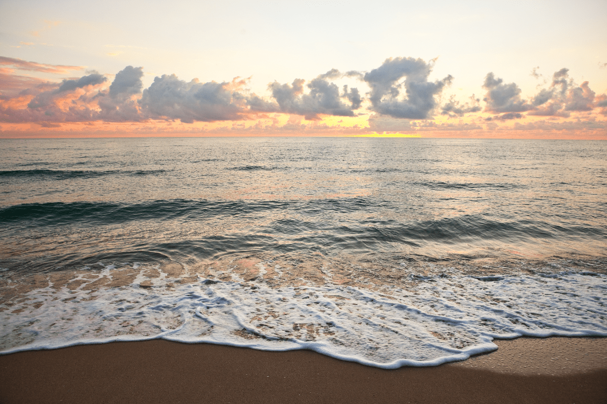 A serene beach scene at sunrise with gentle waves lapping against the shore and a sky filled with scattered clouds illuminated by soft, warm colors.