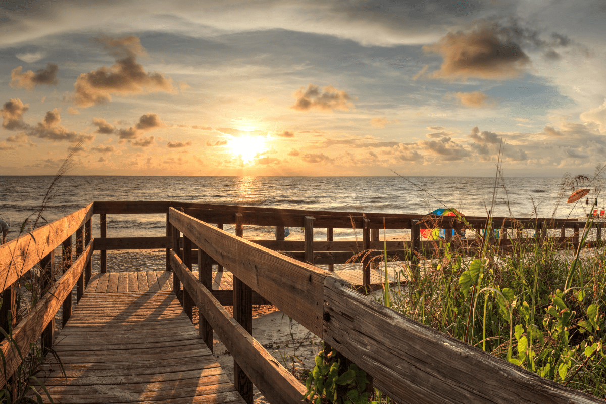 A wooden boardwalk leads to a sandy beach at sunset, with vibrant clouds, gentle waves, and lush greenery on the sides.