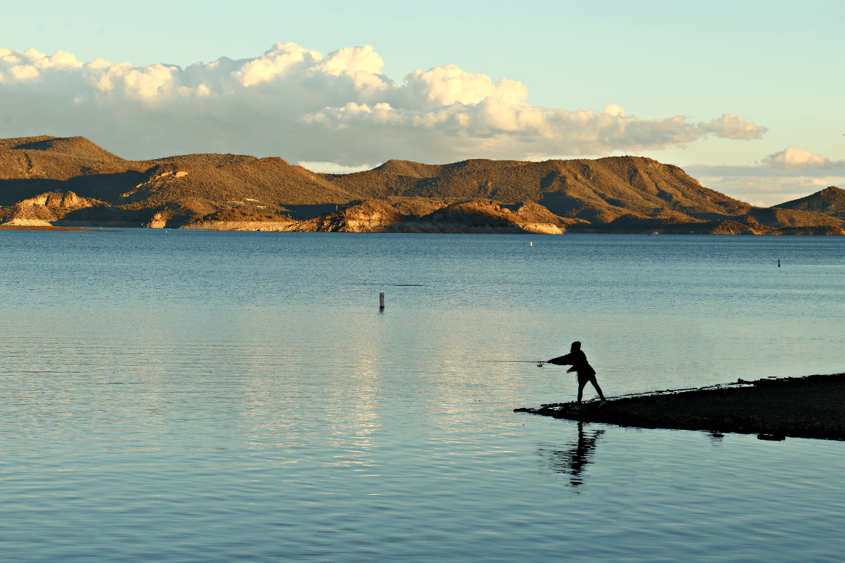 A person is fishing at the edge of a calm lake with mountainous terrain and scattered clouds in the background during sunset.