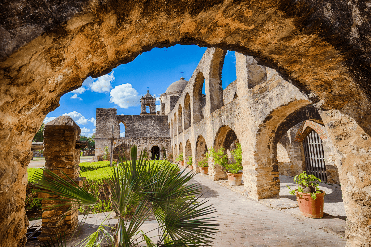 A historic stone archway frames a view of a sunlit courtyard with potted plants, leading to an ancient church with domed towers under a bright blue sky.