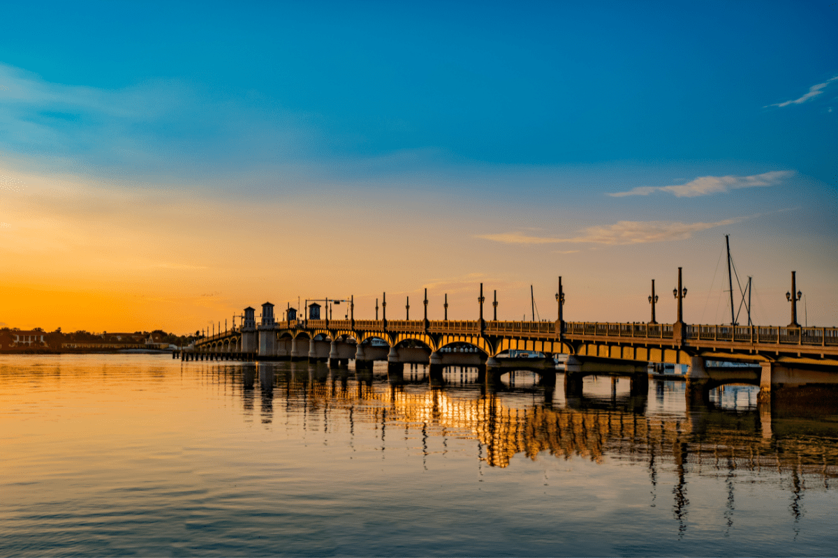 A picturesque view of a long bridge with ornate lampposts spans over calm water at sunset, reflecting warm orange and blue hues in the sky and water.
