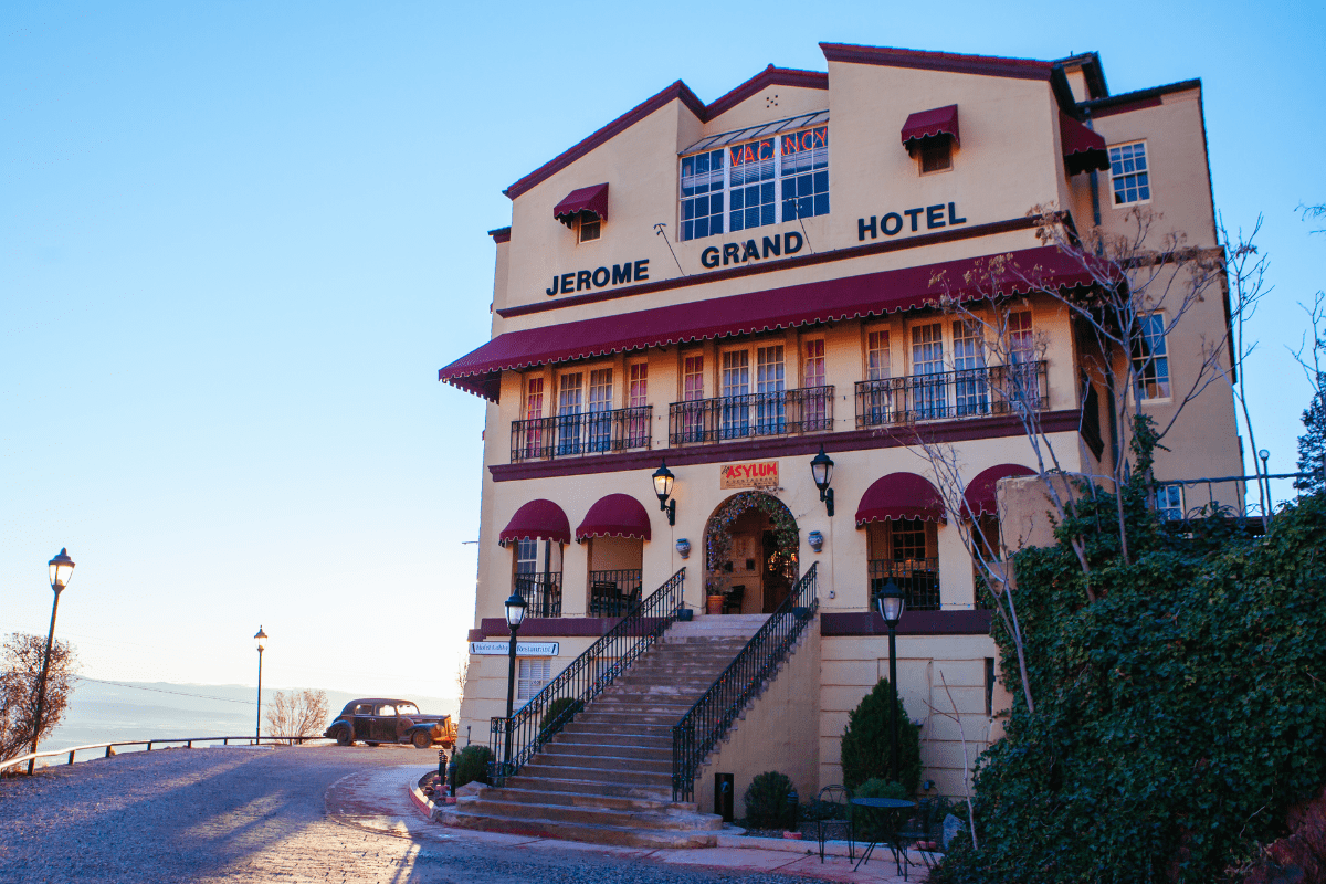 The image shows the historic Jerome Grand Hotel, a large beige building with red awnings and a sign for the Asylum restaurant, set against a clear blue sky.
