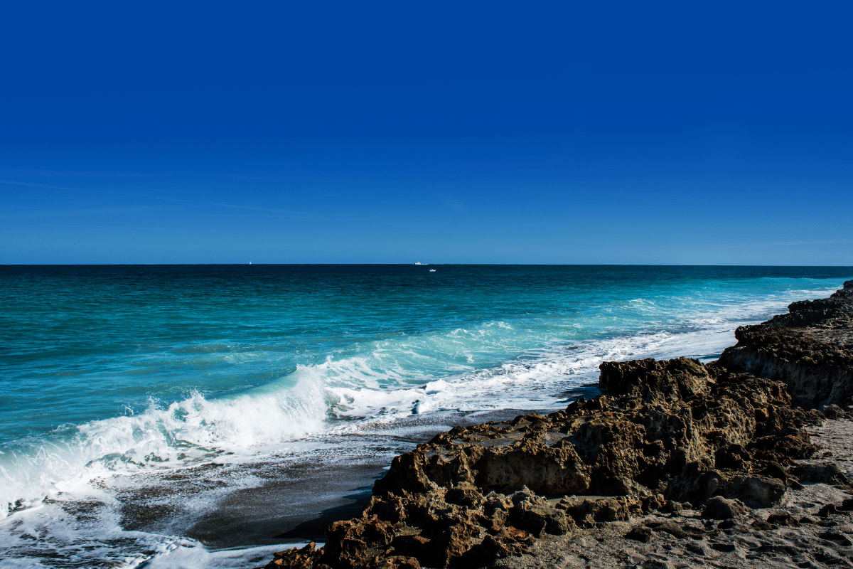 A rocky shoreline with waves crashing against the rocks under a clear blue sky and a distant horizon.