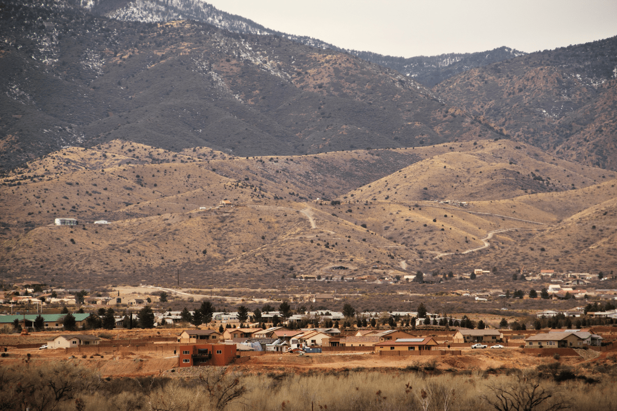 A small rural community with scattered houses is set against a backdrop of rolling hills and rugged mountains under an overcast sky.