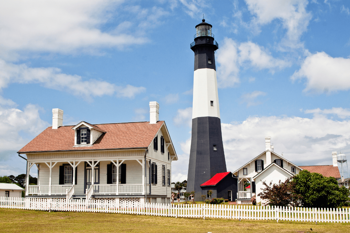 A tall black and white lighthouse stands beside a charming white house with a brown roof, surrounded by a white picket fence under a partly cloudy sky.