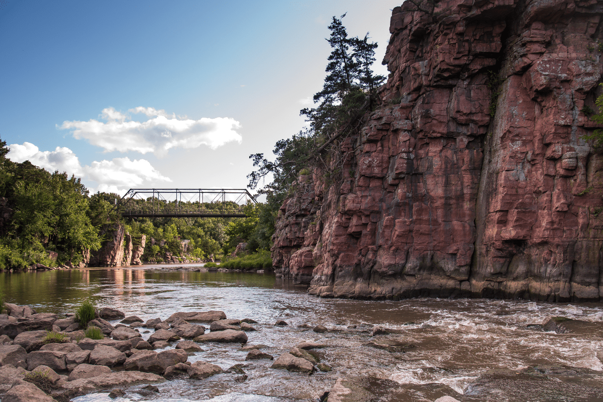 A scenic view of a rocky river with a metal bridge spanning between red cliffs, surrounded by lush greenery under a partly cloudy sky.