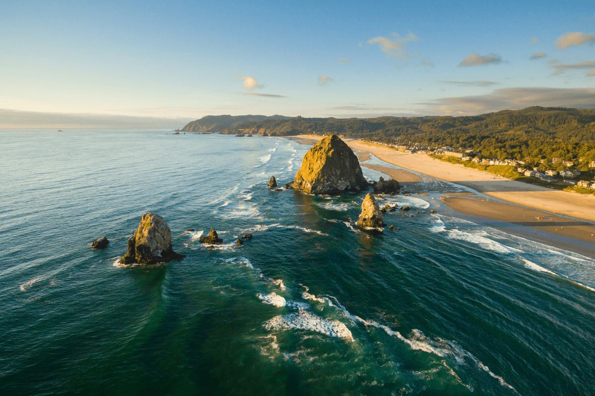 Aerial view of a coastal landscape featuring large rock formations in the ocean, a sandy beach, and a forested area under a clear blue sky.