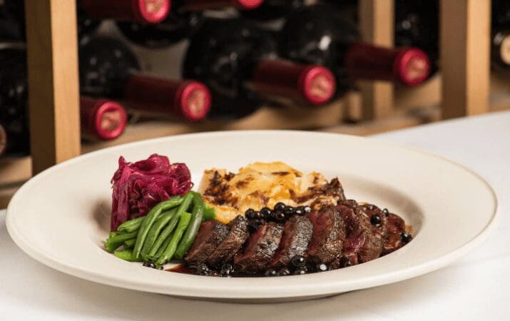 Plate of sliced steak with sauce, mashed potatoes, green beans, and red cabbage; wine bottles in background.