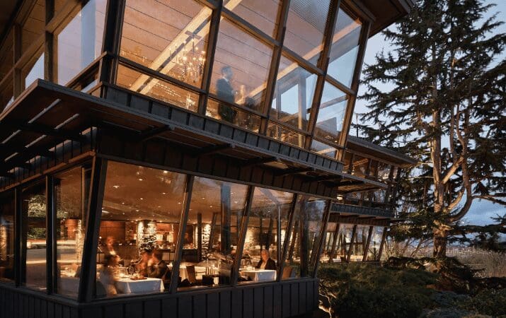 A modern, glass-walled restaurant with people dining inside, surrounded by trees at dusk.