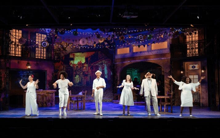 Performers in white outfits sing on a colorful, decorated stage with a festive backdrop.