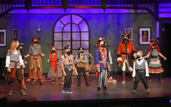 A group of actors in pirate costumes perform on stage with a nautical backdrop.