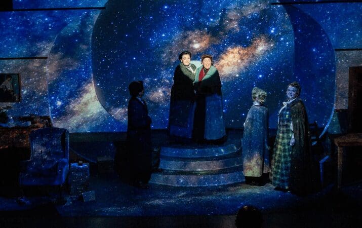 Actors on stage with a starry galaxy backdrop, wearing period costumes, performing a dramatic scene.