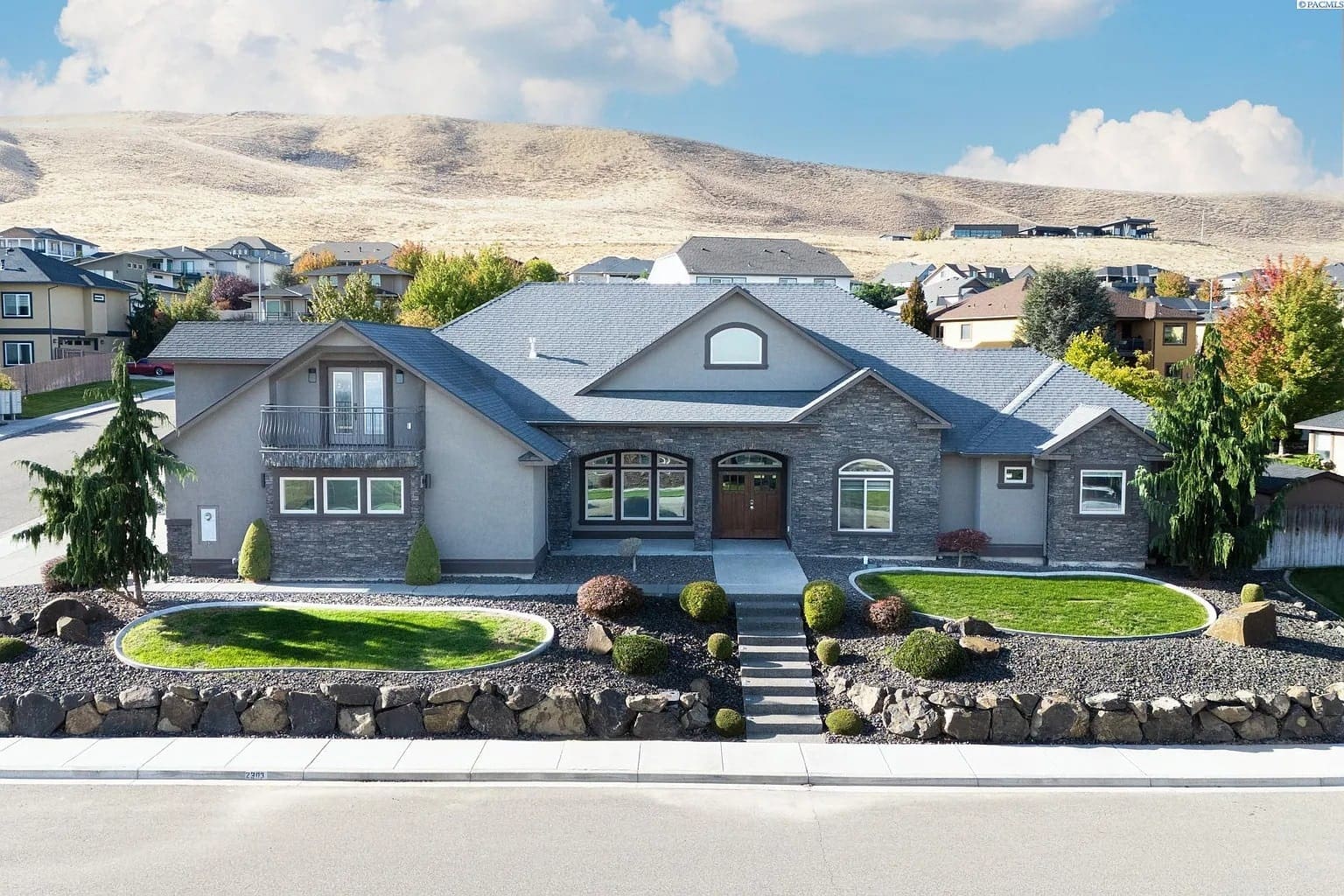 Single-story modern house with gray exterior, manicured lawn, and stone landscaping, set against hilly background.