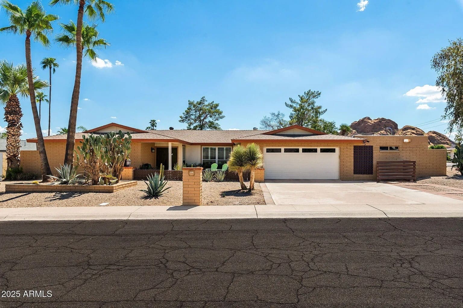 Single-story house with desert landscaping, palm trees, and a two-car garage under a clear sky.