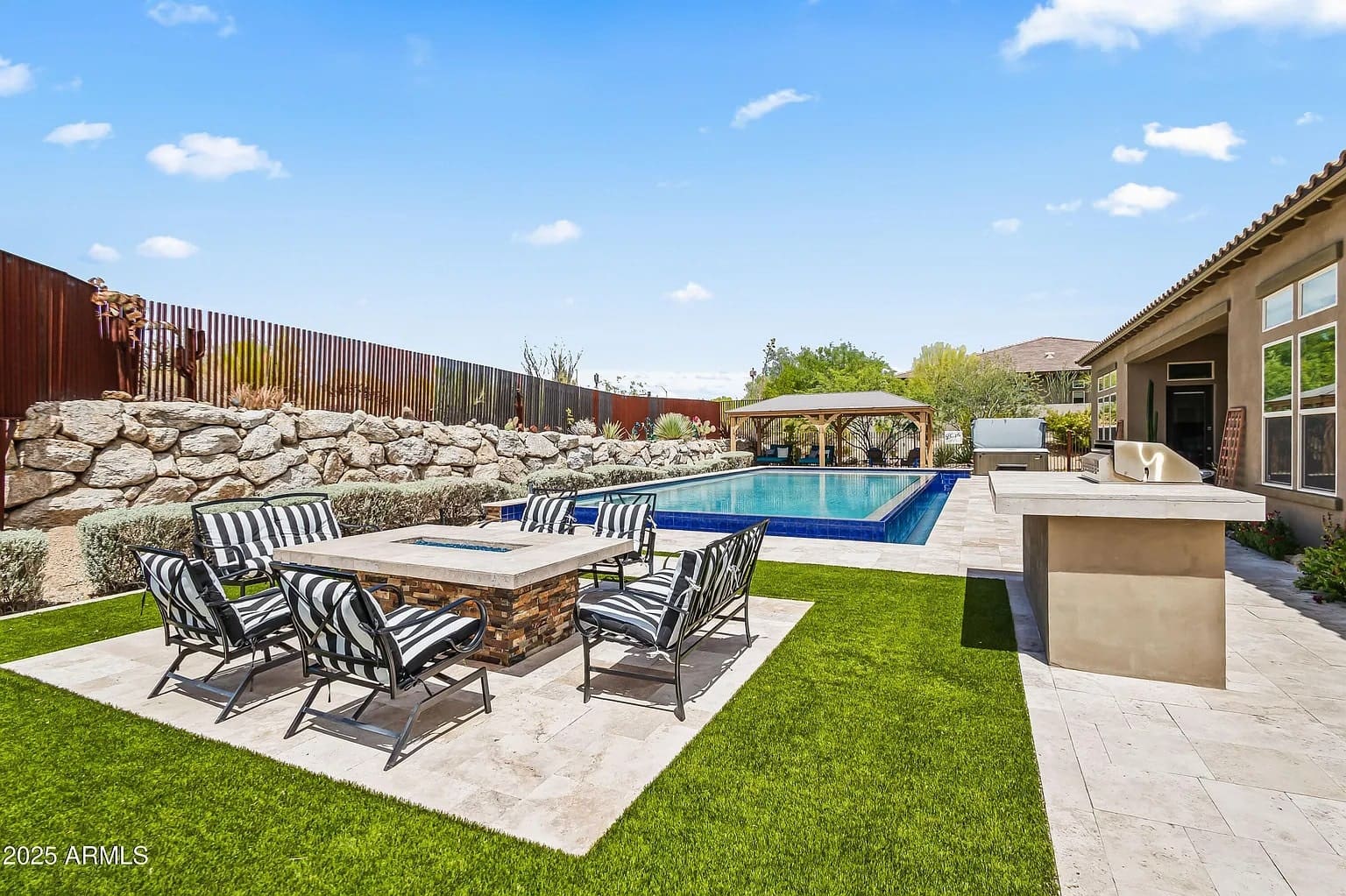 Backyard with a pool, patio furniture, fire pit, and outdoor kitchen under a clear sky.