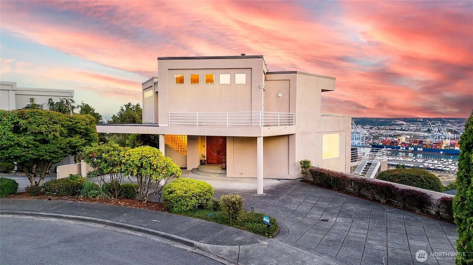 Modern house with large windows, surrounded by trees, overlooking a cityscape at sunset.