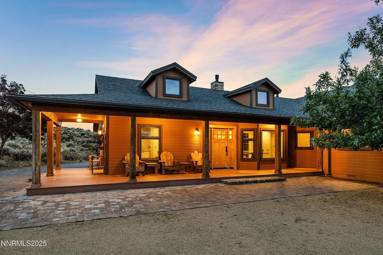 A rustic wooden house with a porch, lit warmly at dusk, surrounded by natural landscape.