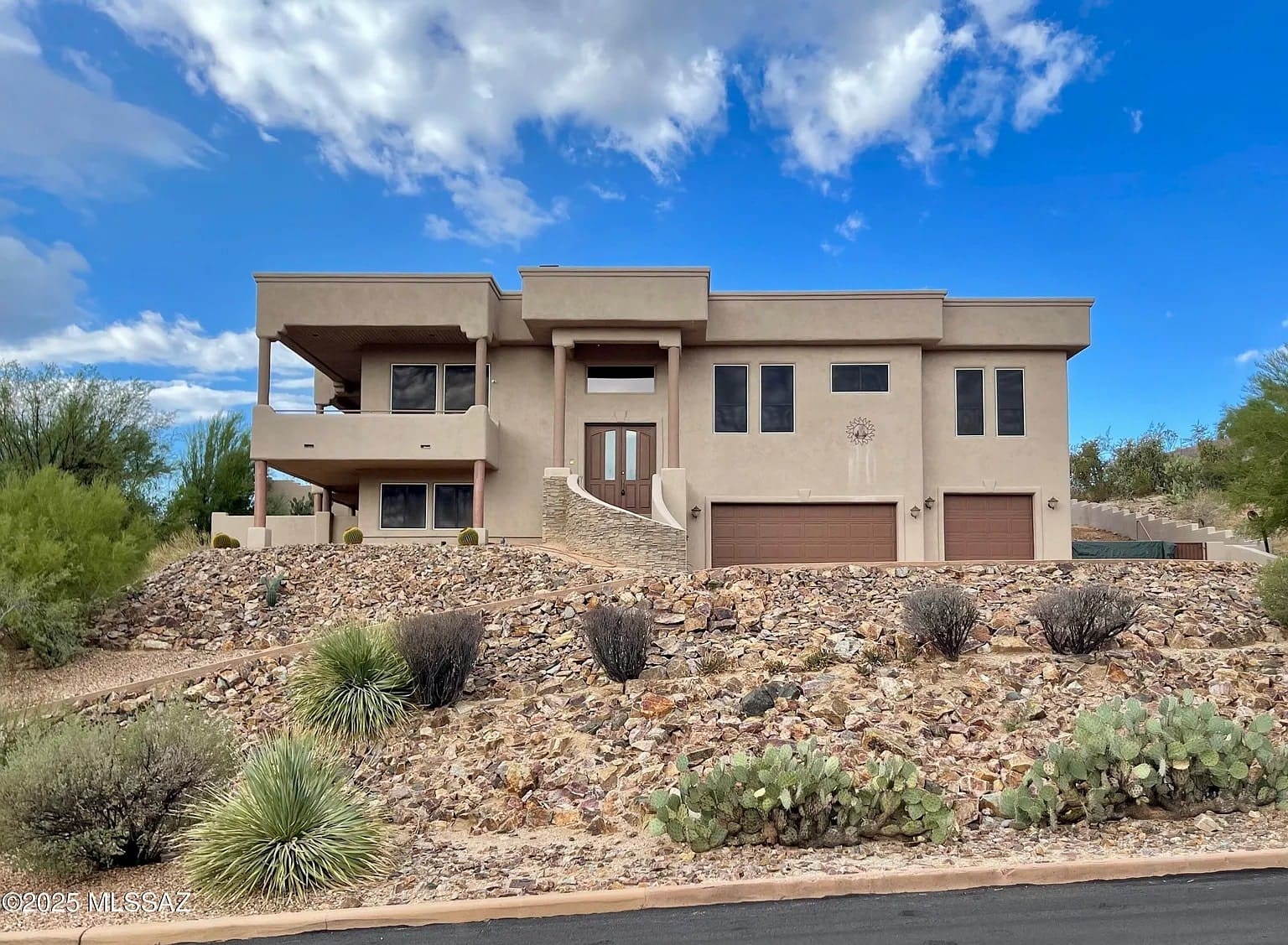 Modern beige house with large windows, two garages, and desert landscaping under a blue sky.