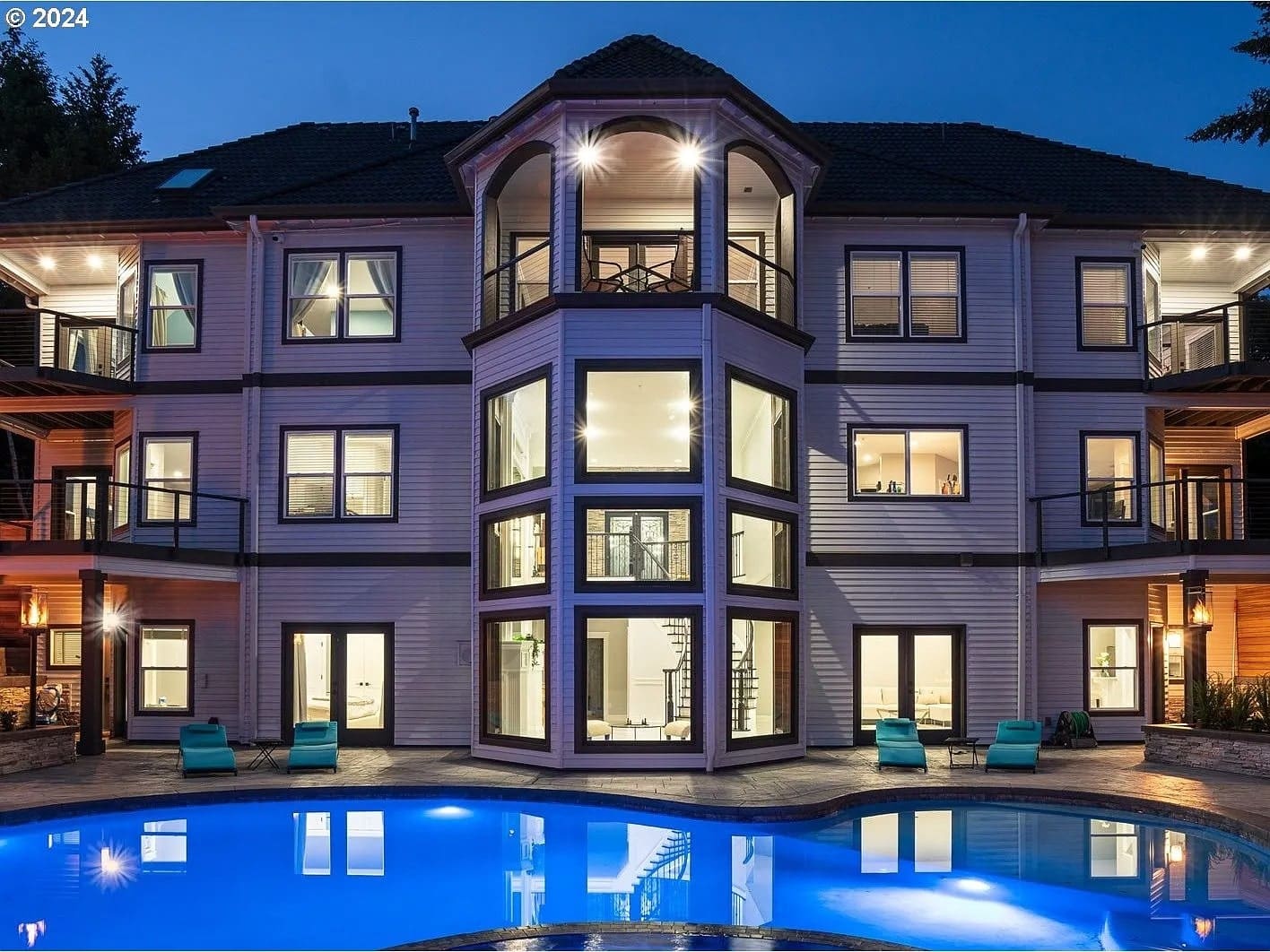 Large, modern house with multiple windows and balconies, overlooking a lit swimming pool at dusk.