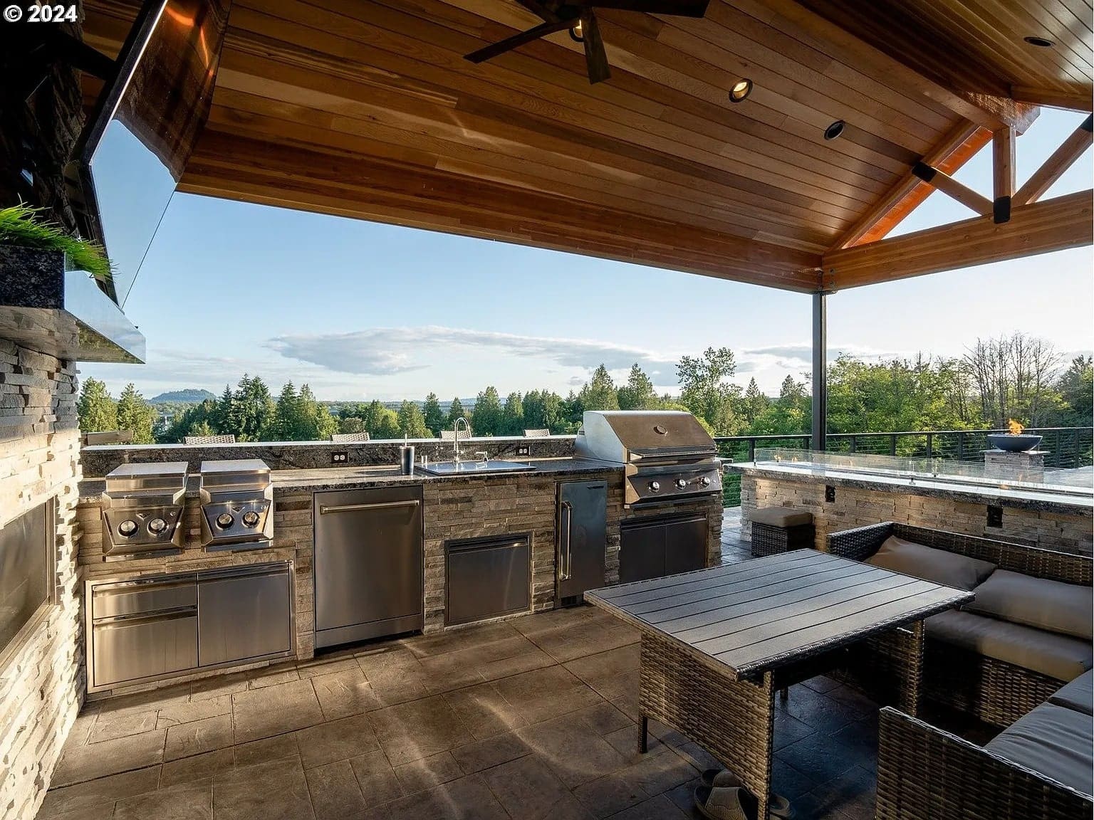 Outdoor kitchen with grills, seating area, and scenic view of trees and distant hills.
