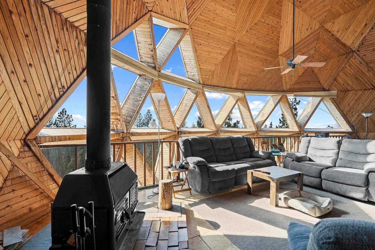 Geodesic dome interior with large windows, cozy seating, wood stove, and ceiling fan.