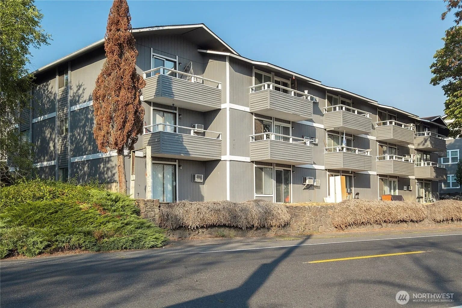 Three-story gray apartment building with balconies, surrounded by trees and bushes, viewed from the street.