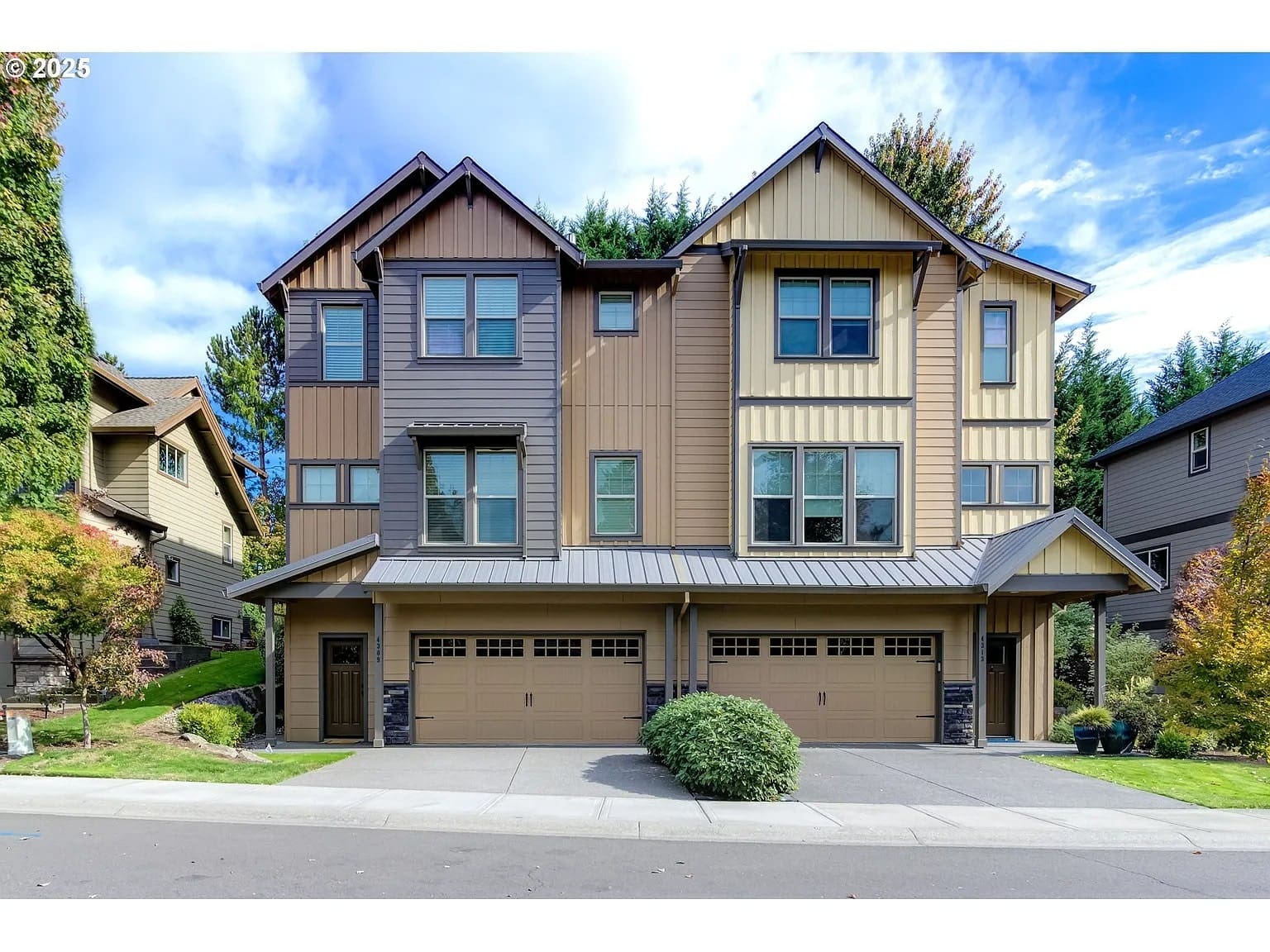 Three-story townhouse with beige and brown siding, three garage doors, and surrounding greenery.