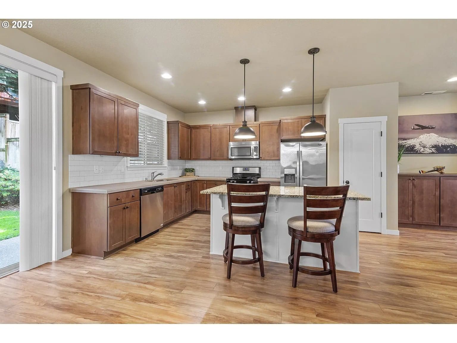 Modern kitchen with wooden cabinets, island with two chairs, stainless steel appliances, and pendant lighting.
