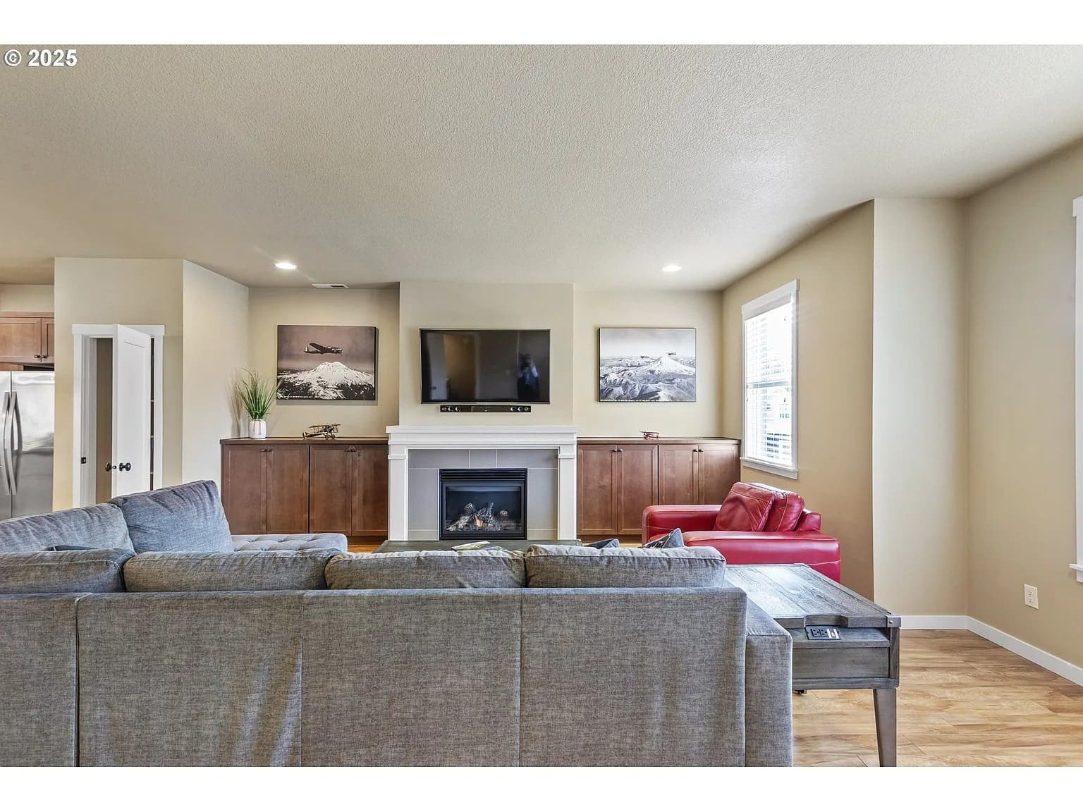 Living room with gray sofa, red chair, TV above fireplace, and landscape photos on walls.