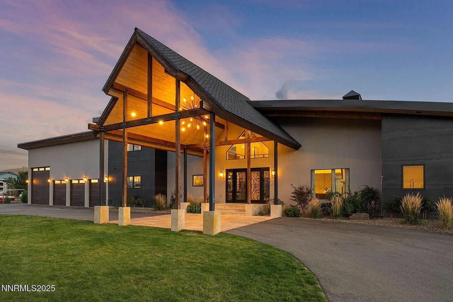 Modern house with large wooden entrance, lit by warm lights, under a colorful evening sky.