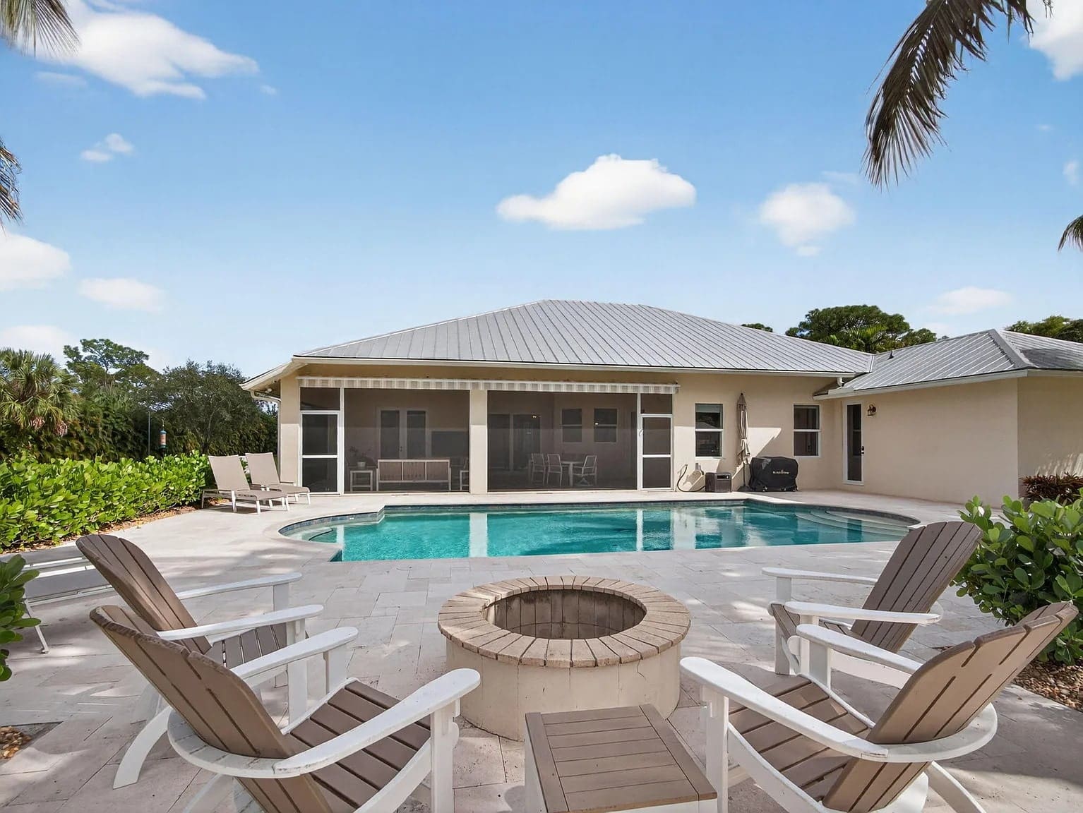 Backyard with a pool, fire pit, and patio chairs, surrounded by greenery under a blue sky.